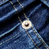 Close-up of dark blue denim with yellow stitching. A copper rivet, marked "S.F. & CO. L.S.", is visible, a testament to the durability of these jeans.