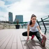 Kobieta wykonuje dynamiczny stretching na drewnianym tarasie nad rzeką, w tle miejski krajobraz z mostem.