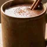 Eine dampfende Tasse vegane heiße Schokolade mit Zimtstange und Kakaopulver auf einem Holztisch.