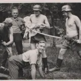 Fünf Männer, einige mit Helmen, posieren mit Waffen. Ein Mann hält eine Puppe. Die Szene erinnert an die Verbrechen der Wehrmacht.