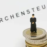 Miniatur-Priester steht auf Münzstapel. Der Schriftzug "Kirchensteuer" deutet auf die Verwendung der Gelder hin.