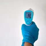 Ręka w niebieskiej rękawiczce trzyma termometr bezdotykowy, pokazujący 36.0°C. Idealne miejsce, gdzie mierzyć temperaturę na czole lub skroni.