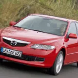 Czerwona Mazda 6 GG w ruchu, z uśmiechniętą kobietą za kierownicą, na tle zieleni.