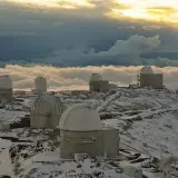 Obserwatoria astronomiczne na ośnieżonym szczycie, otoczone morzem chmur. W oddali widać złote światło zachodzącego słońca, a na pierwszym planie "la silla" - jedno z kopułowych budynków.