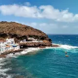 Pueblo costero blanco anidado en la ladera rocosa de **Tufia**. El mar azul turquesa choca contra las rocas, con pequeñas embarcaciones flotando.