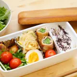 A delicious rice bento box with pork rolls, chicken meatballs, a hard-boiled egg, cherry tomatoes, and a side salad.