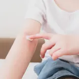 Dziecko smaruje kremem zaczerwienioną skórę na ramieniu, łagodząc objawy azs u dzieci.