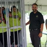 Smiles abound at a jail and bail fundraiser. A woman in a "Jail and Bail" shirt stands by a cage with women in neon shirts. A deputy and a woman holding a baby smile nearby.