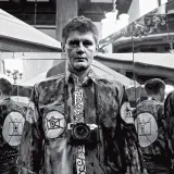 Krzysztof Miller w lustrzanym odbiciu, z aparatem na szyi, w koszuli z motywem oka i aparatu.