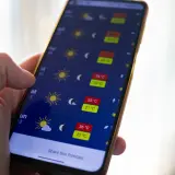 Dłoń trzyma smartfon z aplikacją pogoda, pokazującą wysokie temperatury i słoneczną aurę.