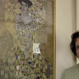 Eine ältere Frau steht neben Gustav Klimts "Adele Bloch-Bauer I", einem goldenen Porträt mit geometrischen Mustern.
