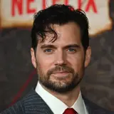 Henry Cavill w garniturze i czerwonym krawacie, z zarostem i niebieskimi oczami.
