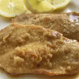 Deliziose scaloppine al limone, succulente e dorate, servite con spicchi di limone fresco su un piatto bianco.