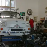 Biały Fiat 600 na podnośniku w warsztacie, gotowy do napraw. W tle narzędzia i motocykle, a nad nimi lampa zapewniająca oświetlenie kanału samochodowego.