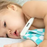 Małe dziecko leży pod kocykiem, a pod pachą ma termometr dla niemowlaka.