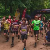 Uczestnicy biegu ultra trail startują w lesie. Widać łuk startowy, flagi i biegaczy w kolorowych koszulkach.