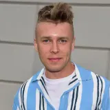 Młody mężczyzna z charakterystyczną fryzurą, ubrany w białą koszulkę i niebiesko-białą koszulę w paski. Czy to właśnie Skolim?
