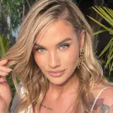 Karolina Gilon z blond włosami, niebieskimi oczami i tatuażem na ramieniu, na tle roślinności.