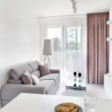 Jasny, bardzo mały salon w bloku z szarą kanapą, stolikiem kawowym i widokiem na balkon. Obok widać łóżko.