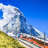 An orange cogwheel train ascends a grassy slope towards the majestic Matterhorn, a perfect scene for hiking in Switzerland.