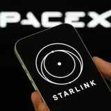 Dłoń trzyma smartfon z logo Starlink na ekranie, w tle logo SpaceX.