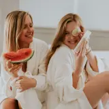 Dwie kobiety w szlafrokach cieszą się chwilą relaksu. Jedna trzyma kawałek arbuza, druga zaś plasterek limonki i buteleczkę z kosmetykiem zawierającym ascorbyl palmitate.