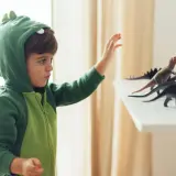 Chłopiec w zielonym stroju dinozaura DIY sięga po figurki prehistorycznych gadów.