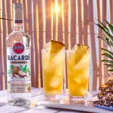 Butelka Bacardi Coconut obok dwóch drinków z ananasem i cała ananasowa piramidka. Idealne na tropikalne przyjęcie!