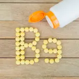 Żółte tabletki ułożone w napis "B9" obok otwartego białego pojemnika na drewnianym tle.