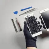 Ręce w rękawiczkach trzymają dwa telefony: jeden z pękniętym ekranem, drugi sprawny. Obok narzędzia do naprawy. Upadek telefonu skutki widoczne.
