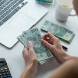 Kobieta liczy pieniądze, sprawdzając, czy kwota wolna od zajęcia jest wystarczająca. Obok leży laptop, okulary i kalkulator.