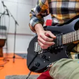 Ręce muzyka grające na czarnej gitarze elektrycznej. W tle stojaki z mikrofonami i bęben. To może być początek nauki, jak zmienić struny w gitarze elektrycznej.