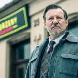 Mężczyzna w eleganckim swetrze i płaszczu, z siwą brodą, stoi przed budynkiem z neonem. To jeden z aktorów z obsady "Minuta ciszy".