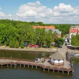 Idealne **Mazury na weekend**: drewniane molo z altaną, jezioro, zielony park i urokliwe miasteczko z czerwonymi dachami.