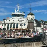 Tętniący życiem deptak w Oslo, z ludźmi siedzącymi w kawiarniach, na tle białego statku i zabytkowej wieży zegarowej. Oslo czy warto? Zdecydowanie tak!