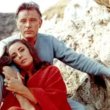 Elizabeth Taylor, żona Richarda Burtona, opiera głowę na jego ramieniu, otulona czerwoną narzutą, siedząc na kamieniach.