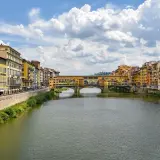 Il Ponte Vecchio, con le sue botteghe colorate, si specchia sull'Arno. Un canoista solitario solca le acque sotto il cielo azzurro e le nuvole bianche.