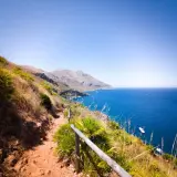 A scenic hiking trail winds through the rugged landscape of Lo Zingaro Nature Reserve, overlooking the brilliant blue Mediterranean Sea dotted with boats.