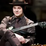 Jack White w brązowym kapeluszu i garniturze gra na gitarze.
