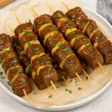 Plusieurs brochettes de bœuf et fromage, garnies de persil frais, sont présentées sur une assiette.