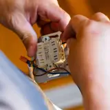 Ręce elektryka podłączają przewody do mechanizmu gniazdka. To kluczowy etap prac, wpływający na koszt instalacji elektrycznej.