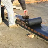 Pracownik nakłada abizol na ceglaną ścianę, przygotowując ją do dalszych prac budowlanych.