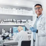 Uśmiechnięty technik farmacji w okularach ochronnych trzyma tablet w laboratorium, gdzie na półkach stoją liczne butelki, a w tle pracuje inna osoba.