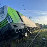 Zielony pociąg towarowy wykoleił się, przechylając się na bok na torach. W tle błękitne niebo i linie energetyczne.