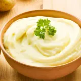 Un tazón de cremoso puré de patatas, decorado con perejil, junto a patatas crudas. ¡Así es como hacer puré de patatas perfecto!