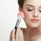 Mujer aplicando rubor en sus mejillas con una brocha. El tono rosado del rubor realza su piel, creando un look natural y radiante.