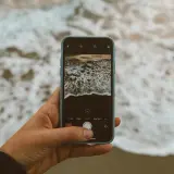 Dłoń trzyma smartfon, którym fotografuje fale. **Expert raw** uchwyci piękno natury.