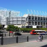 St James' Park w Newcastle, z charakterystyczną białą konstrukcją dachu, otoczony zielenią i ruchliwą ulicą z autobusem i rowerzystą.