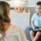 Chłopiec w niebieskiej koszuli siedzi na pufie, rozmawiając z psychoterapeutką.