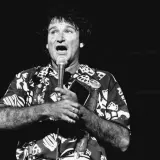 Robin Williams, w hawajskiej koszuli, śpiewa do mikrofonu, trzymając w dłoni notes.